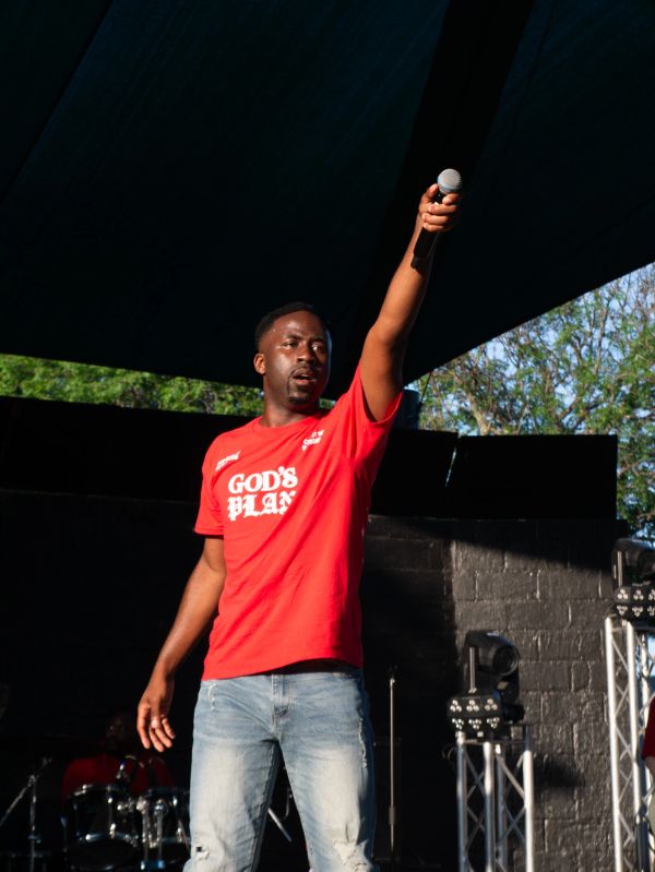 Man holding microphone on stage wearing a red "God's Plan" shirt.