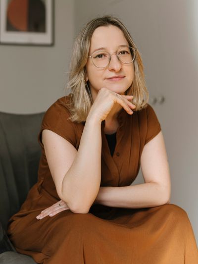 A thoughtful woman wearing glasses and a brown dress sits with her hand resting on her chin.