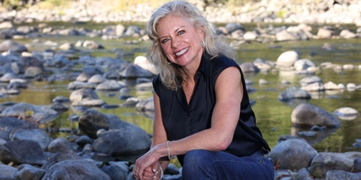 Woman smiling, seated on a rock by a river surrounded by stones and trees.