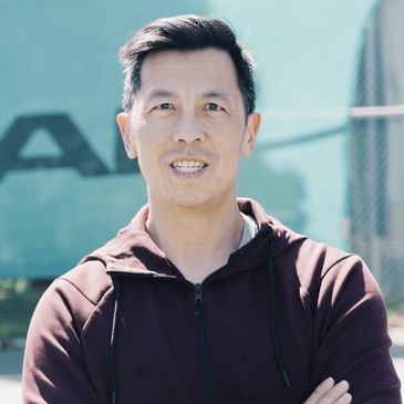 Man in a maroon hoodie smiling outdoors with arms crossed.