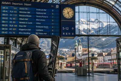 Traveler with skis at a snowy mountain train station looking at the departure board.