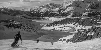 Skiers descending a snowy mountain slope with a vast, rugged mountain range in the background.