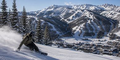Snowboarder carving down a snowy mountain with pine trees and a village below.