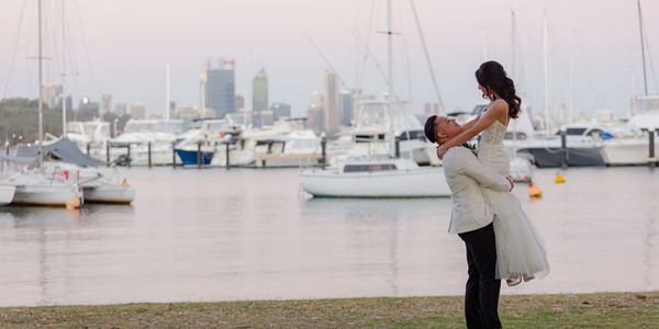 Newlywed couple embraces by the marina with boats and city skyline in the background.