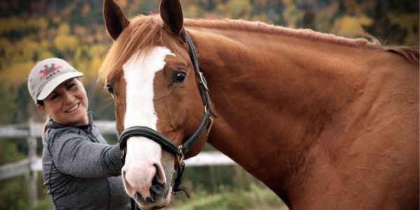 A smiling woman with a horse outdoors in a natural setting.