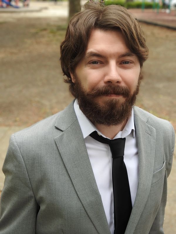 Man with beard in a grey suit and black tie outdoors.