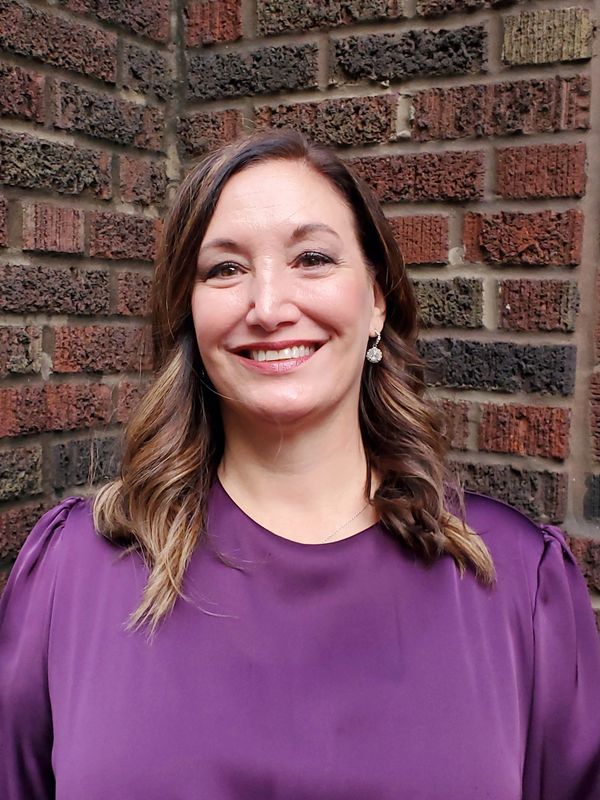 Smiling woman in a purple blouse standing against a brick wall.