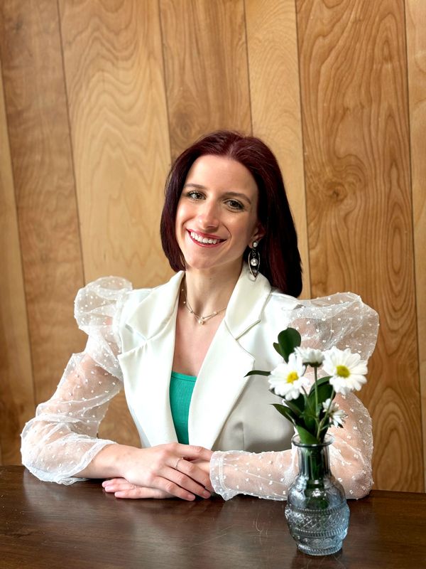 Young woman with red hair smiles, wearing a white blazer with sheer sleeves, sitting at a table with daisies.