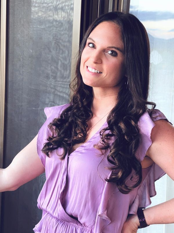 A woman with long curly hair in a lavender dress smiles by a window.