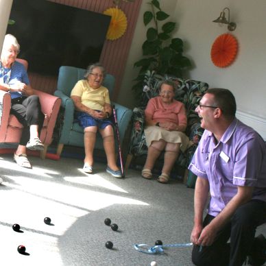 A group of people playing carpet boules with an activity coordinator. They’re sitting in a sunny roo