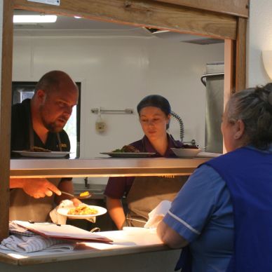Two chefs are dishing up a meal behind a service hatch. A care assistant is waiting to take the food