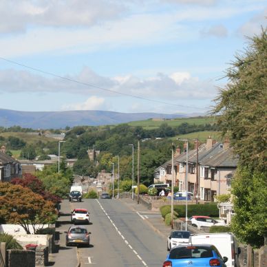 Looking down a road into a town. There are cars parked and you can see trees and hills in the distan
