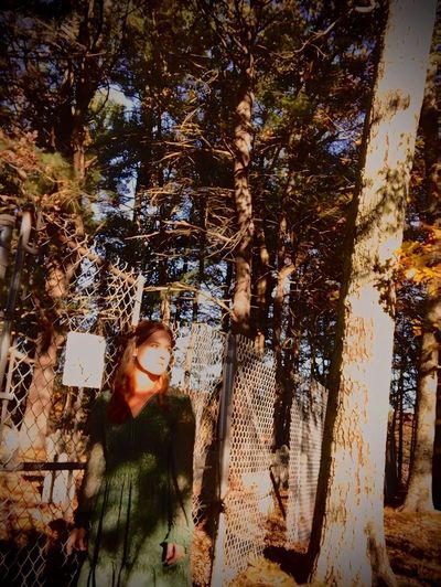 Jessie wearing a green dress standing in front of a chain link fenced in cemetery in the woods