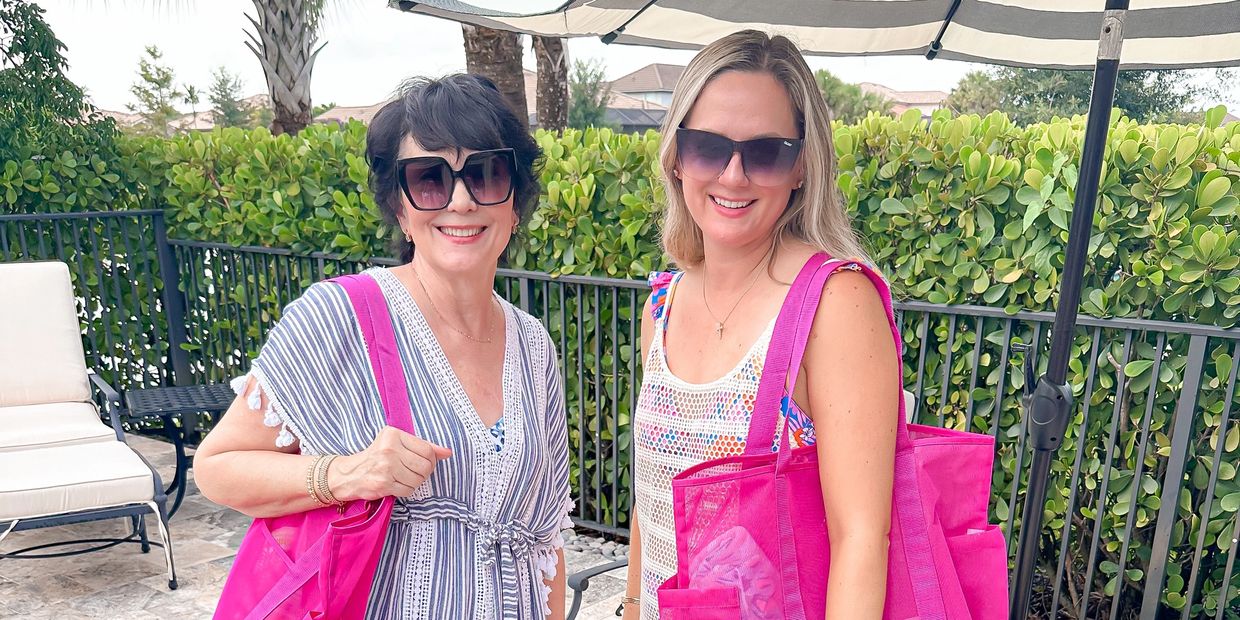 Two women smiling outdoors with large pink tote bags and sunglasses.