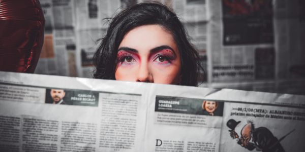 Woman with dramatic makeup peeks over a newspaper with a backdrop of newspaper pages.