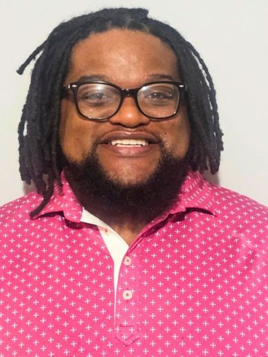 Smiling man with dreadlocks and glasses wearing a pink patterned shirt.