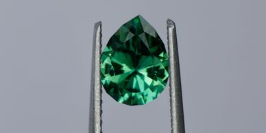 A pear-shaped green gemstone held by tweezers against a gray background.