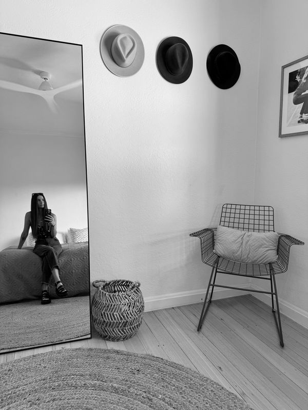 A cozy corner with a green wire chair, hats on the wall, and a large mirror reflecting a woman.