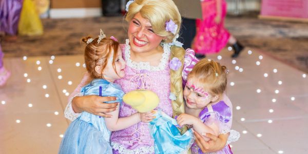 Woman dressed as Rapunzel hugs two smiling little girls in princess costumes.
