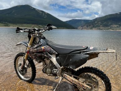 A dirt bike parked at the edge of a lake with mountains in the background.