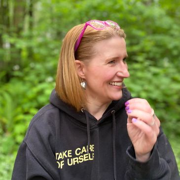 Smiling woman in a hoodie outdoors with greenery background.