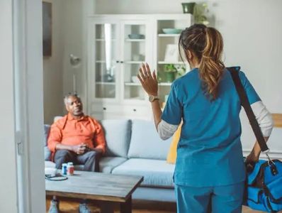 A nurse greets a man sitting on a couch in a home setting.