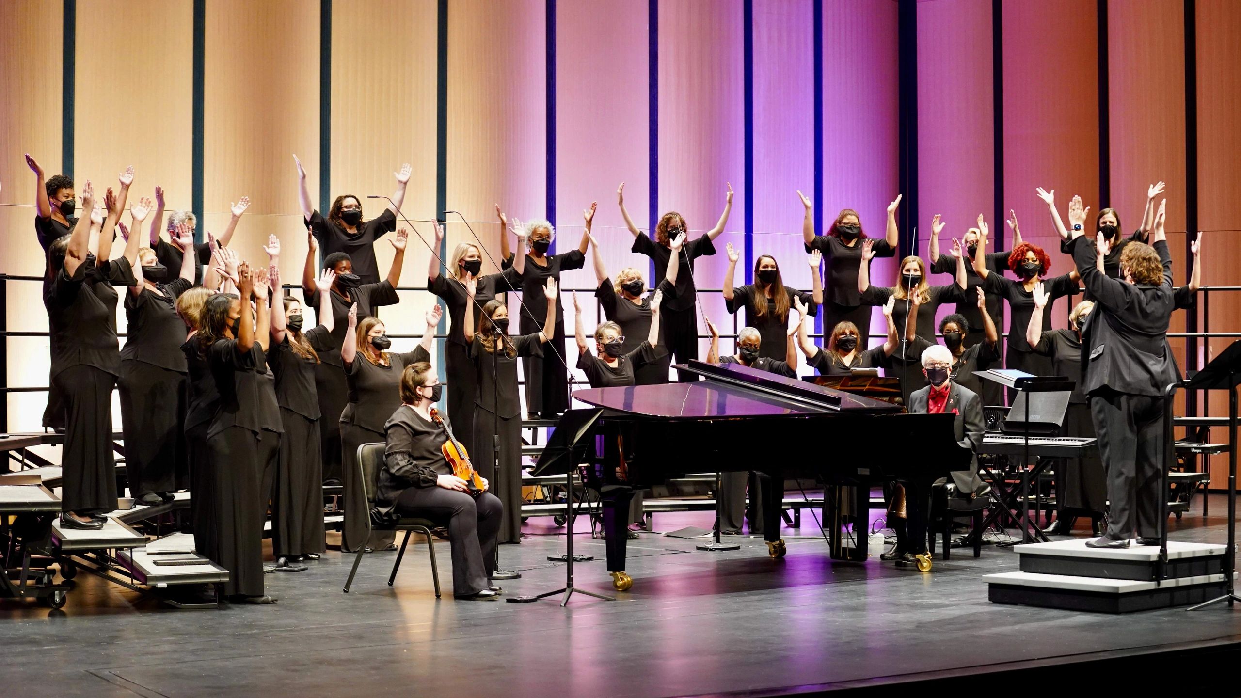 A choir of masked singers perform onstage with their arms lifted, facing a conductor doing the same,