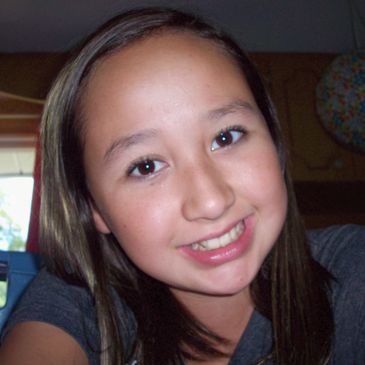 A young girl smiling indoors with long brown hair.