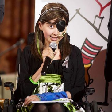 Young girl with an eye patch speaking into a microphone while sitting in a wheelchair.