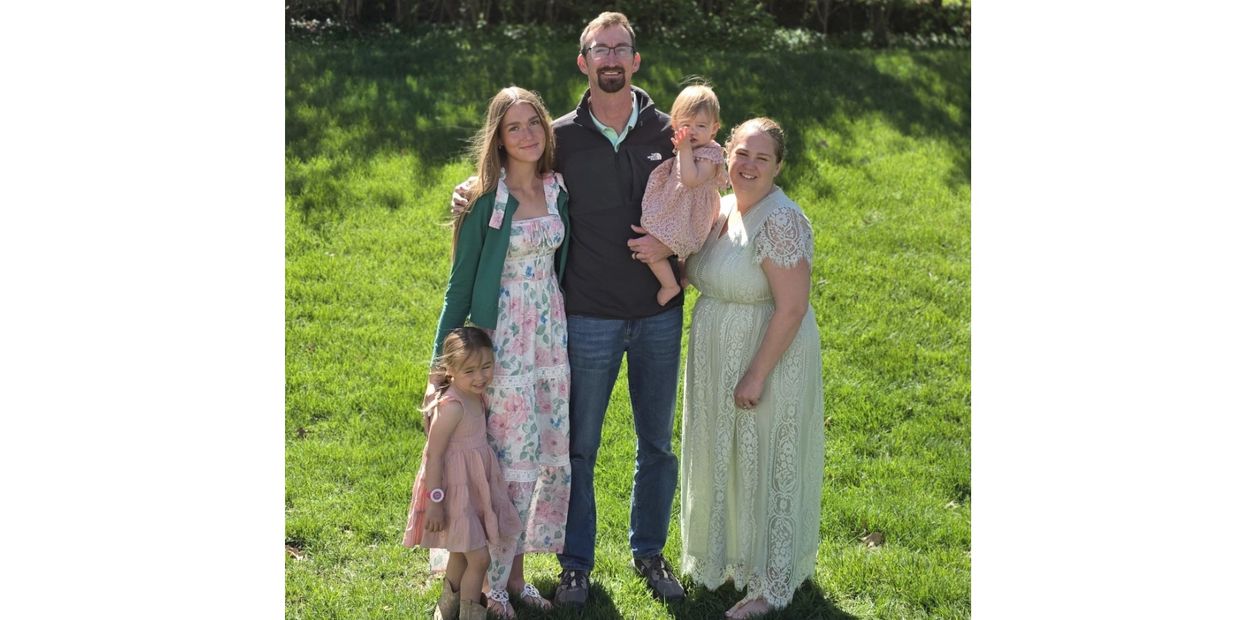 A family of five posing outdoors on a sunny day, smiling at the camera.