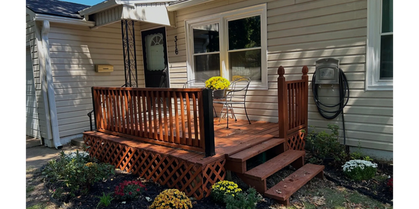 Small wooden porch with two chairs and flower pot in front of beige house.
