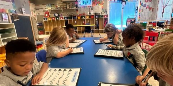Young children practicing writing on dry erase boards at a classroom table.