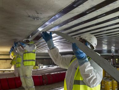 Workers apply adhesive strips to a ceiling in an industrial setting.