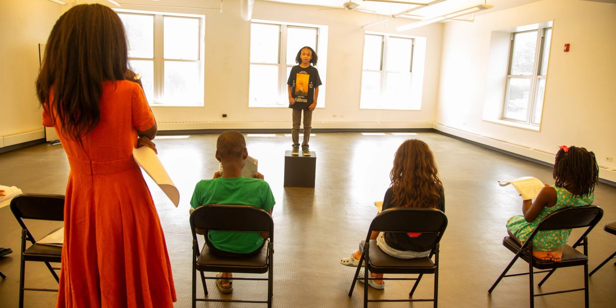 Child performing while others observe with scripts.