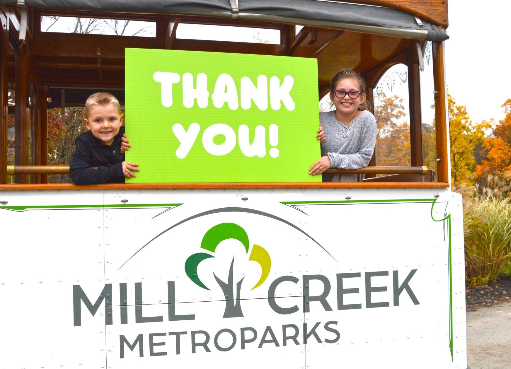 Two kids on a Mill Creek Metroparks trolley holding a bright green thank you sign.