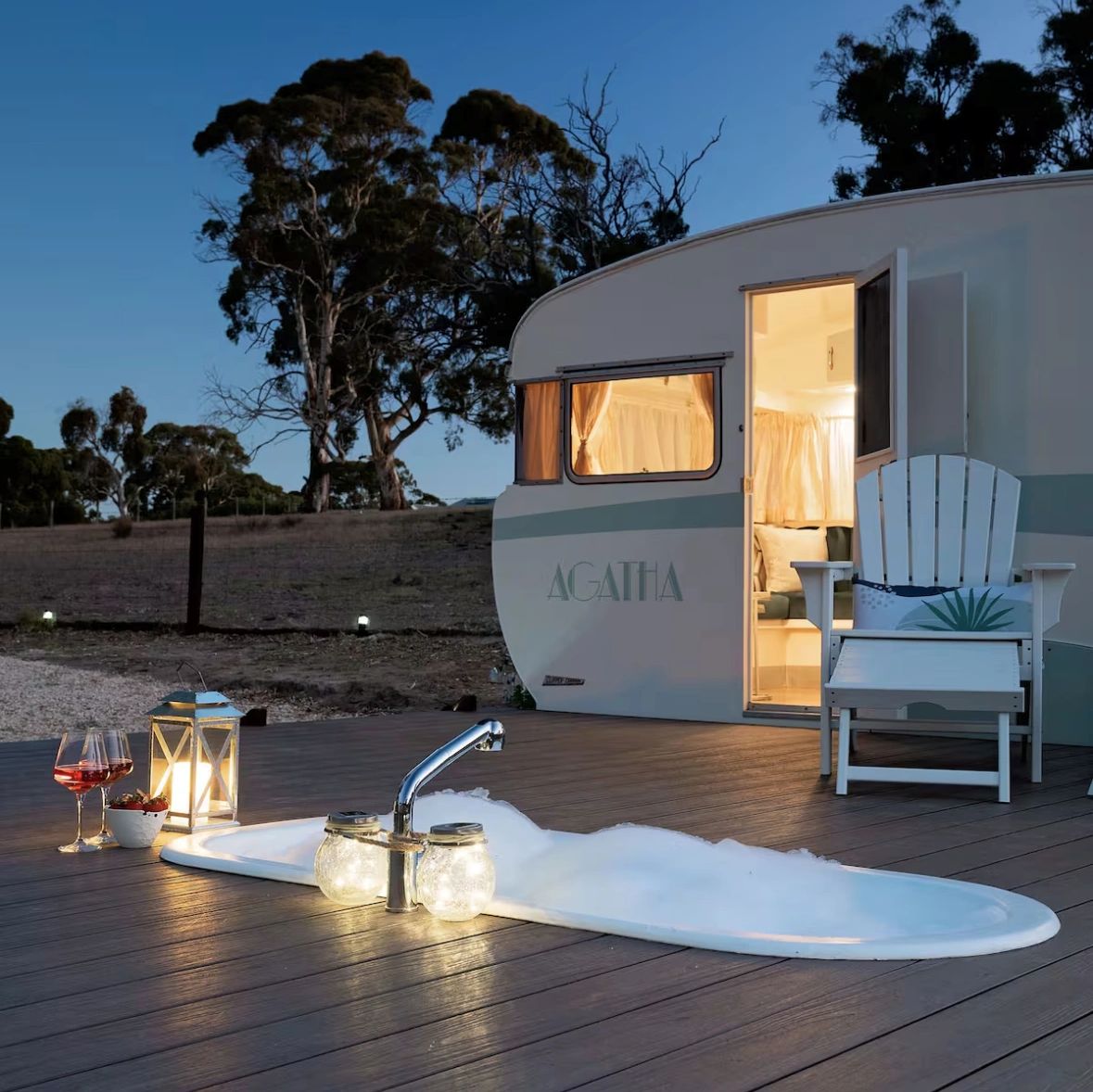 Outdoor bathtub with bubbles beside a cozy caravan at dusk.