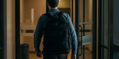 Man with a backpack walking through a glass door in a dimly lit hallway.
