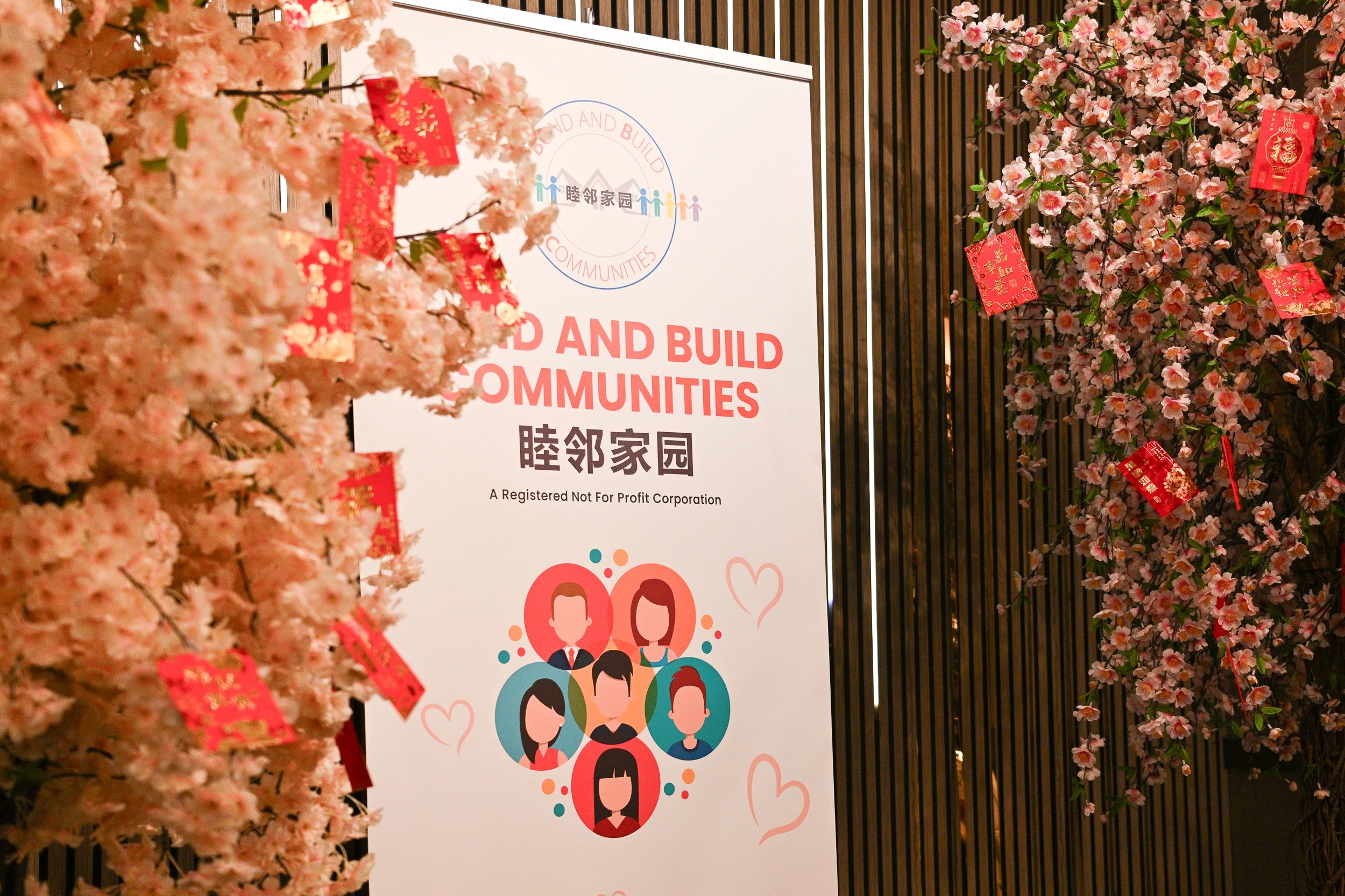 Cherry blossom trees decorated with red envelopes beside a community organization banner.