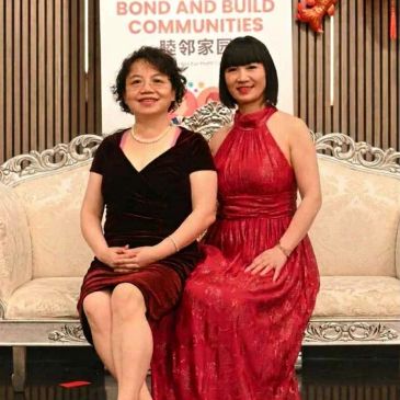 Two women in elegant dresses sitting on a vintage sofa at a community event.