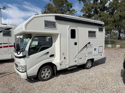 Compact white camper van parked on gravel with trees in the background.