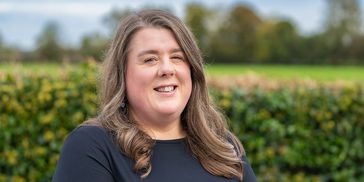 Smiling woman in a black top outdoors with greenery behind her.