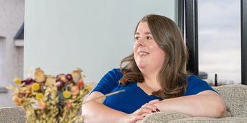 A woman in a blue dress relaxes on a beige couch smiling.