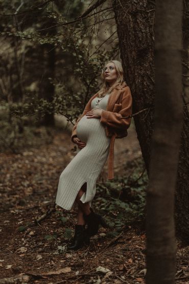 Pregnant woman in a white dress and brown coat leaning against a tree in a forest.