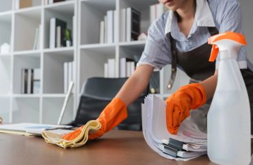 Person cleaning a desk with orange gloves and cleaning spray bottle.
