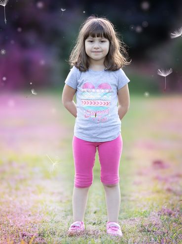 A young girl standing outdoors with a soft smile and hands behind her back.