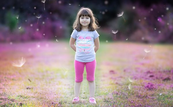 A young girl stands in a field with dandelion seeds floating around her.