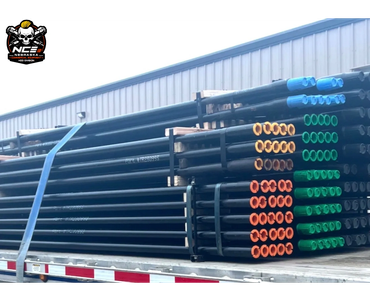 Stacked metal pipes with colored caps loaded on a flatbed trailer near a warehouse.