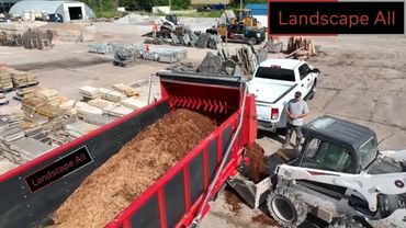 Bobcat loading mulch into a red dump truck at a landscaping yard.