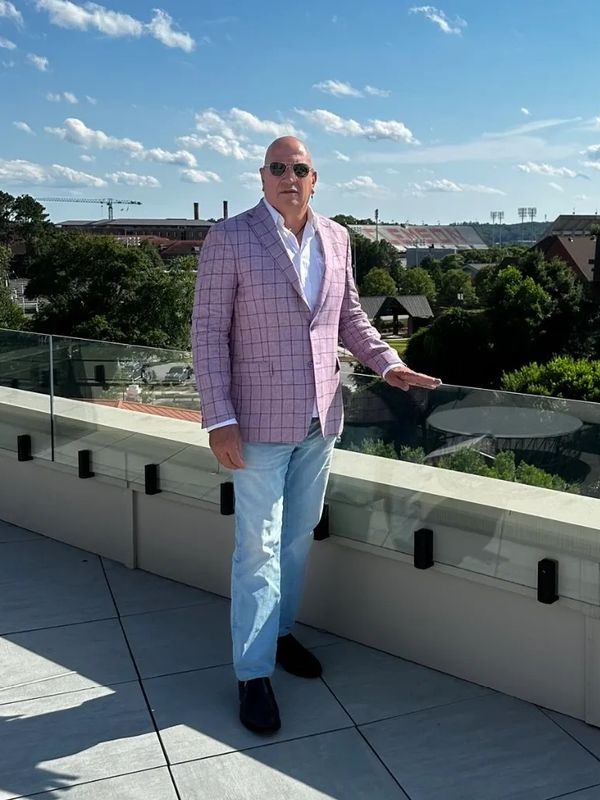 Man in sunglasses and pink checkered blazer on rooftop with city view.