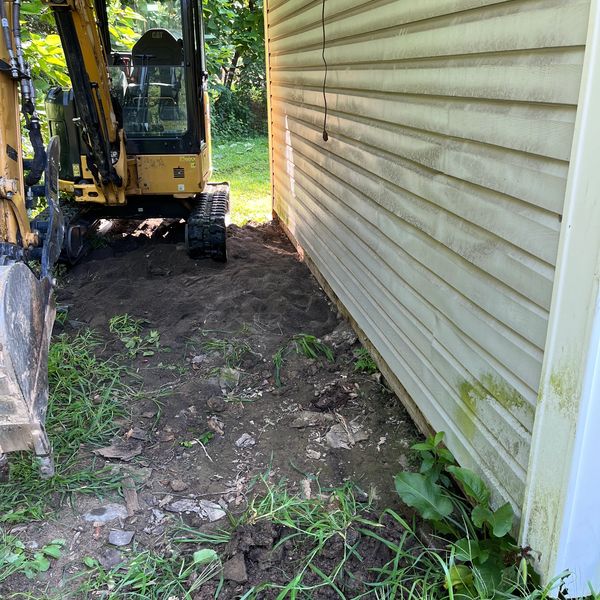 Excavator clearing soil beside a yellow building in a garden area.